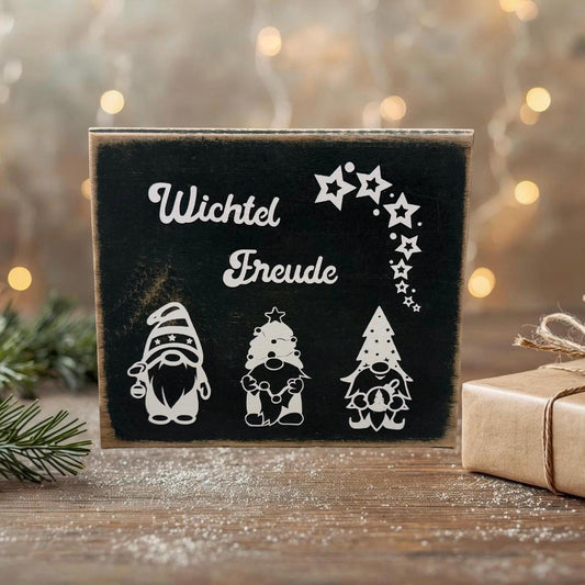 Deko Weihnachtswichtel auf Holz mit dem weißen Schriftzug „Wichtel Freude“ und drei stilisierten Wichtelmotiven auf schwarzem Hintergrund, arrangiert auf einem rustikalen Holztisch mit Tannenzweig und Geschenk im Hintergrund.