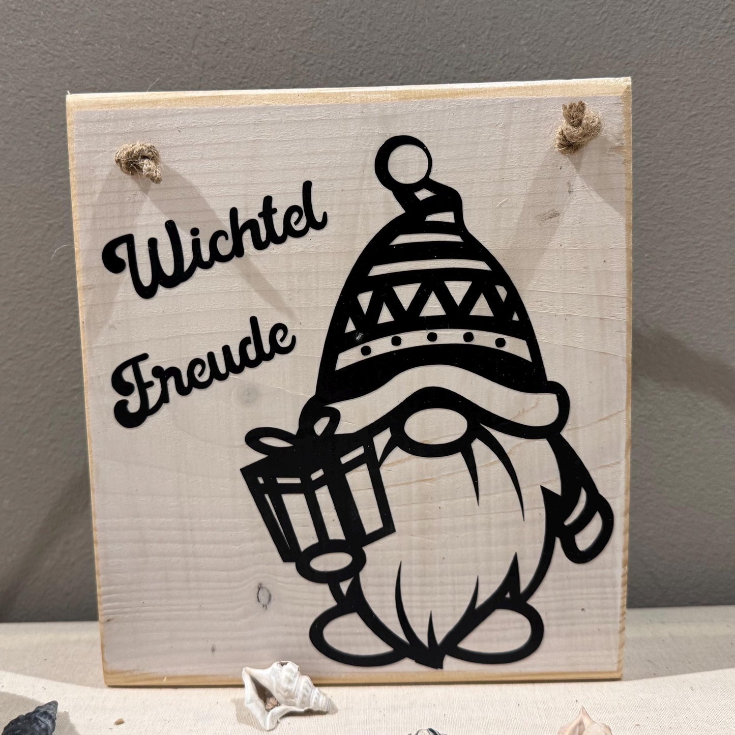 Quadratisches Holzschild mit dem Schriftzug "Wichtel Freude" und einer schwarzen Zwerg-Illustration, die ein Geschenk hält.