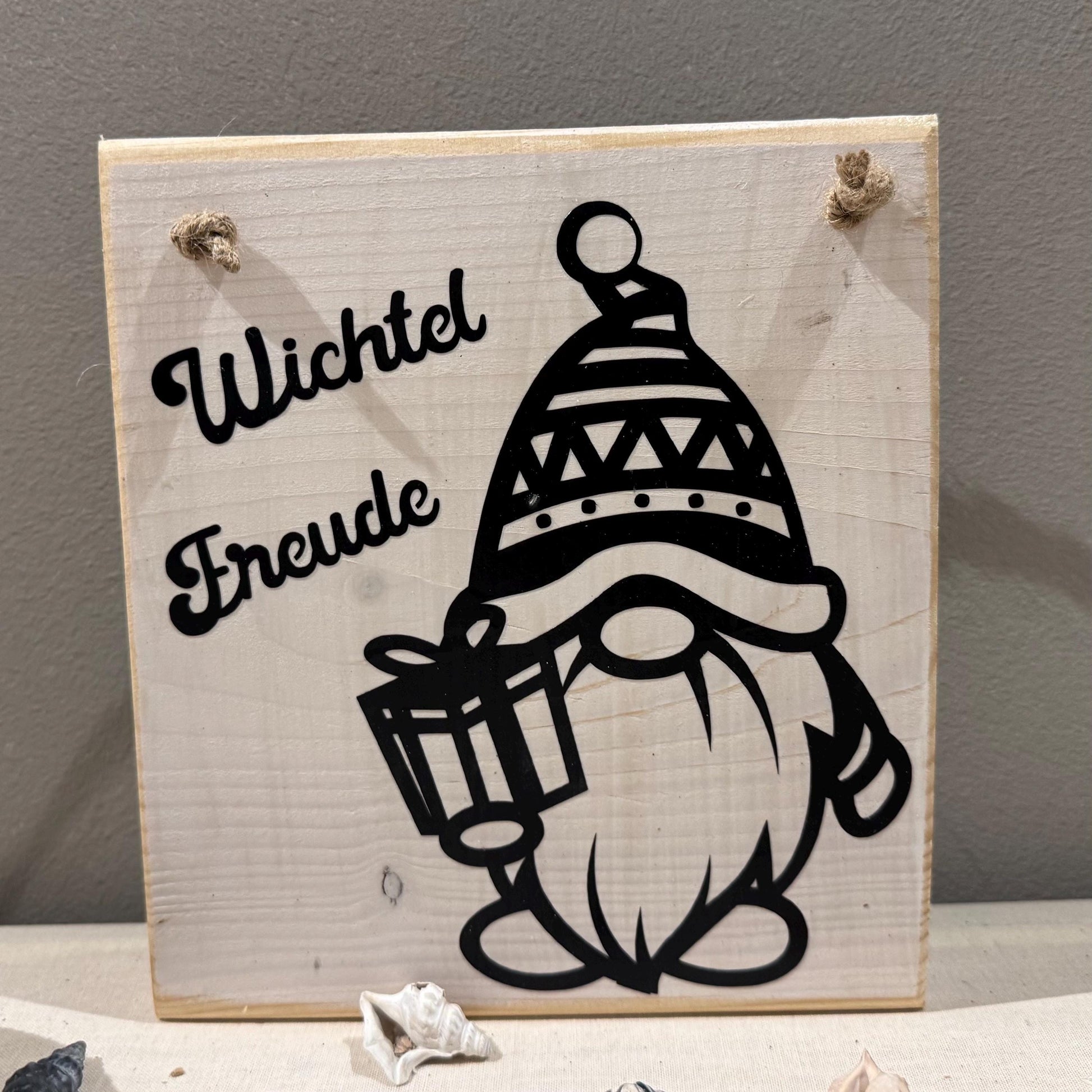 Quadratisches Holzschild mit dem Schriftzug "Wichtel Freude" und einer schwarzen Zwerg-Illustration, die ein Geschenk hält.