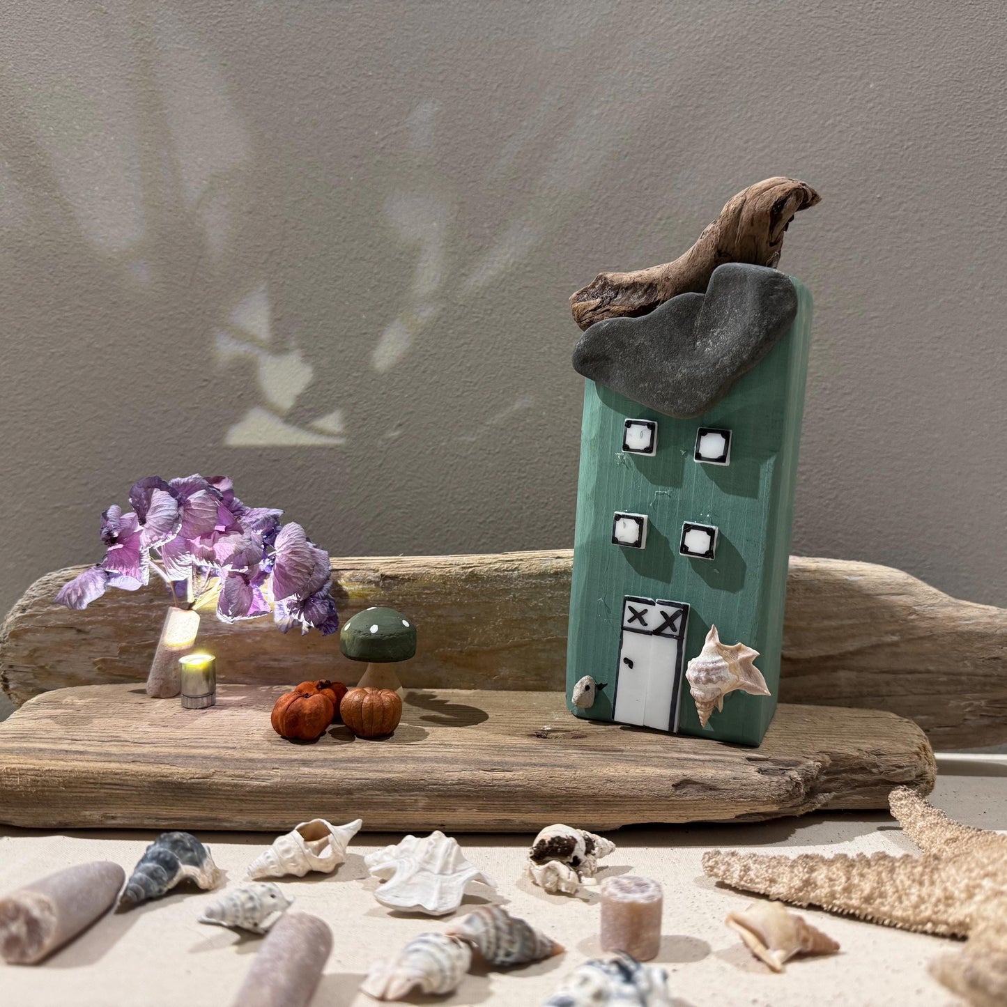 Ein handgefertigtes grünes Holzhaus mit Steindach und Treibholzdetails. Die Szene umfasst Kürbisse, Muscheln und Blumen