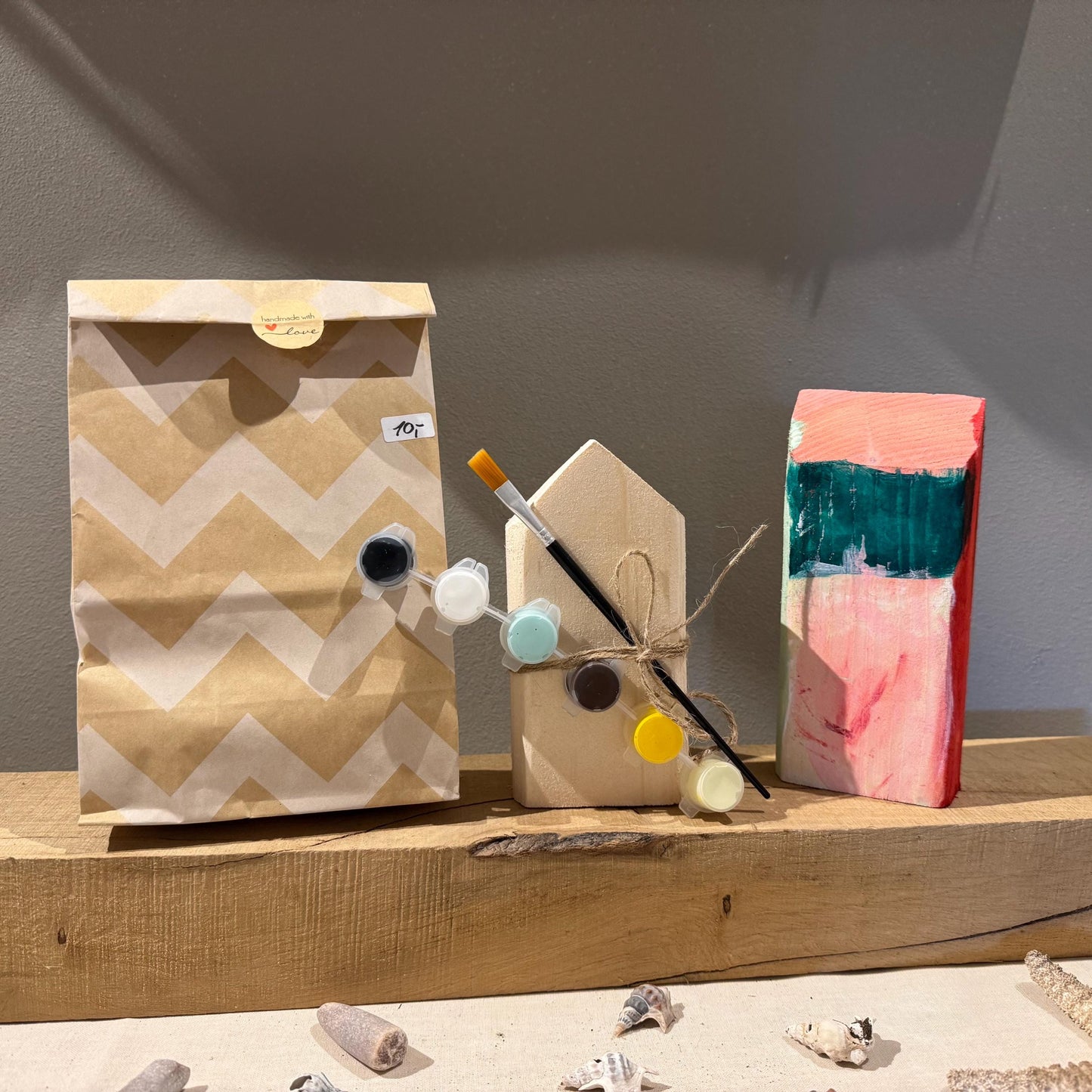 Ein Bastelset mit einem Holzhaus, Farbe und Pinsel. Eine braune Papiertüte mit Zickzackmuster und ein bemalter Holzblock