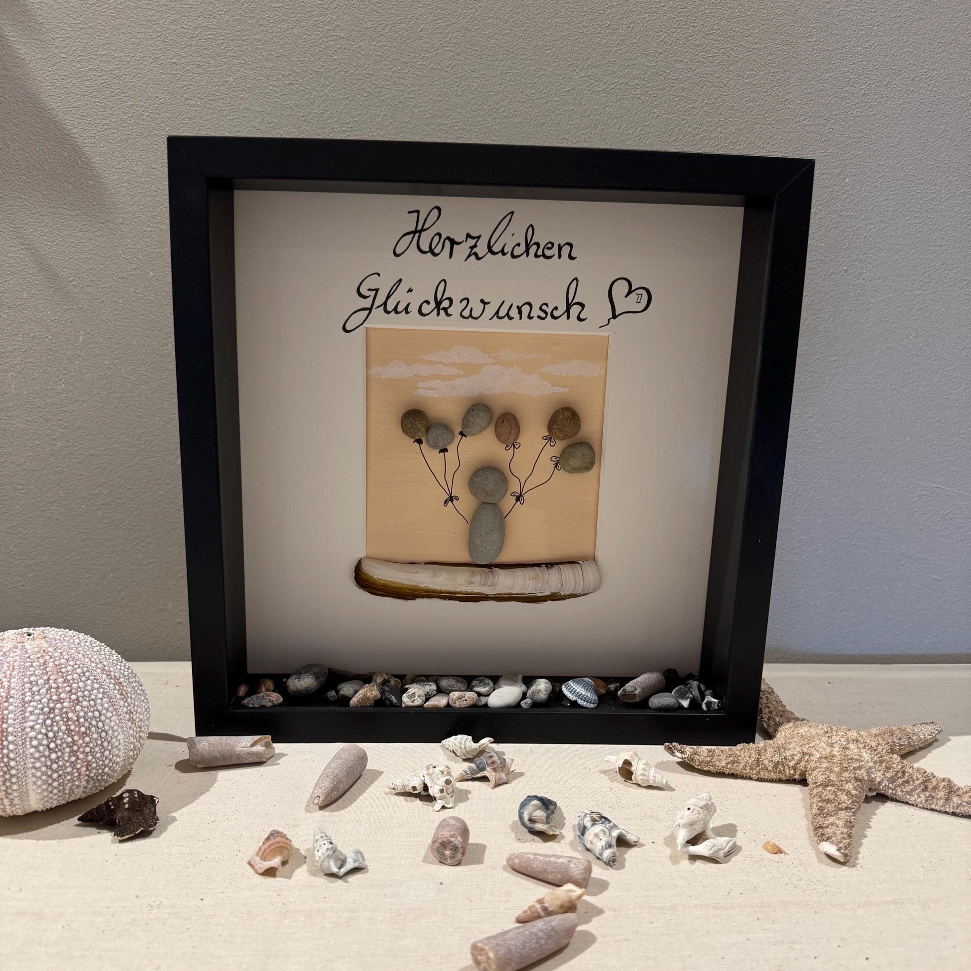 Ein gerahmtes Kunstwerk mit Holzrahmen. Das Kunstwerk zeigt ein Kieselkunst-Design einer Person, die Ballons hält
