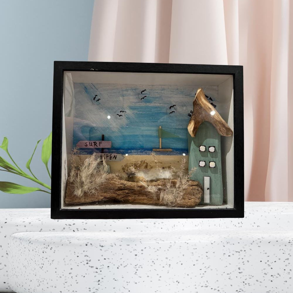 Ein gerahmtes Schattenbox-Diorama mit einer Miniatur-Strandszene. Die Szene umfasst ein kleines Haus von Treibholzzauber79