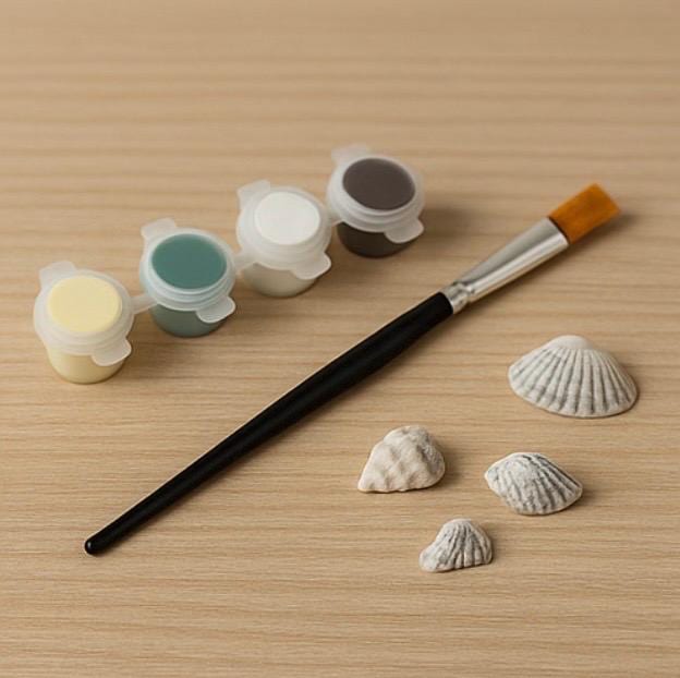 Ein Mal-Set mit vier Farbtöpfen in Gelb, Türkis, Weiß und Grau, einem Pinsel und vier kleinen Muscheln von Treibholzzauber79