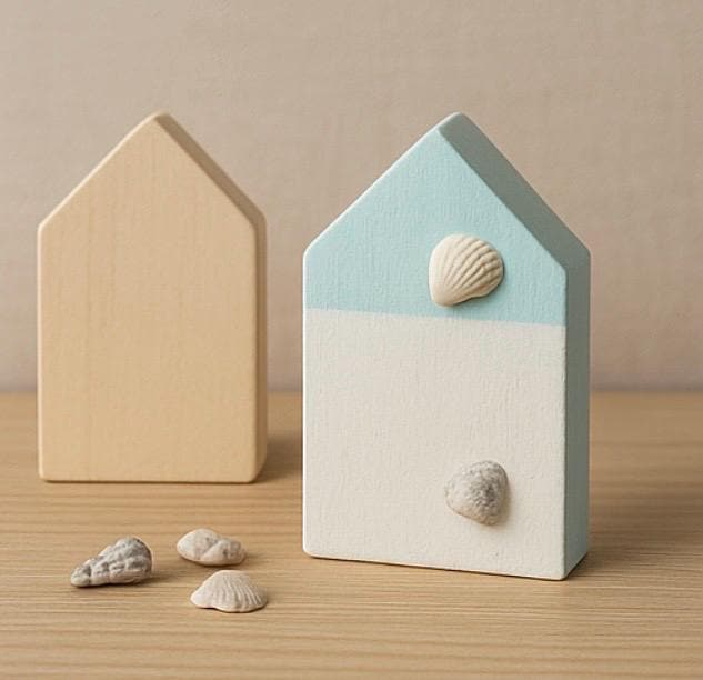 Zwei Holzdekorationen in Hausform. Eine ist aus Naturholz, die andere ist hellblau und weiß bemalt und mit Muscheln versehen.