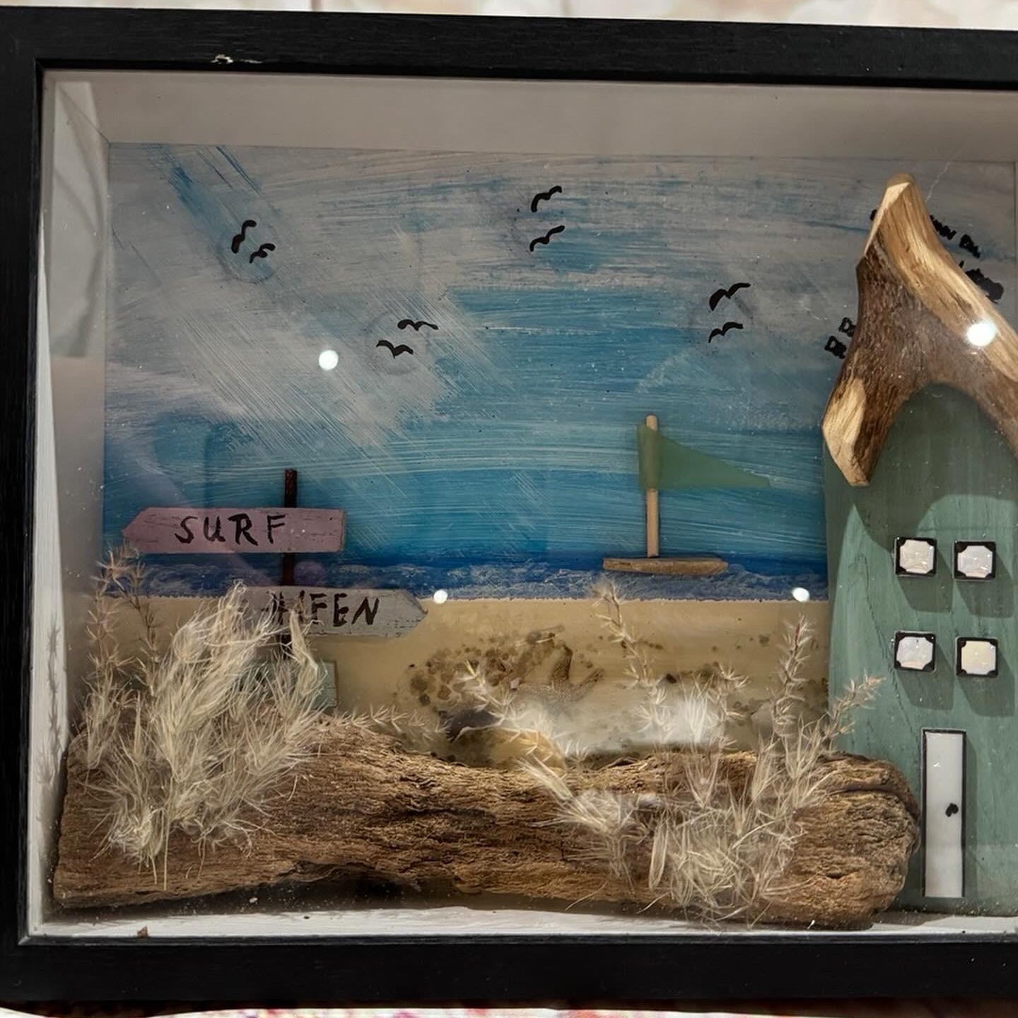Maritimes 3D Strandbild mit Treibholz, Haus und Surf Hafen Wegweiser im schwarzen Bilderrahmen