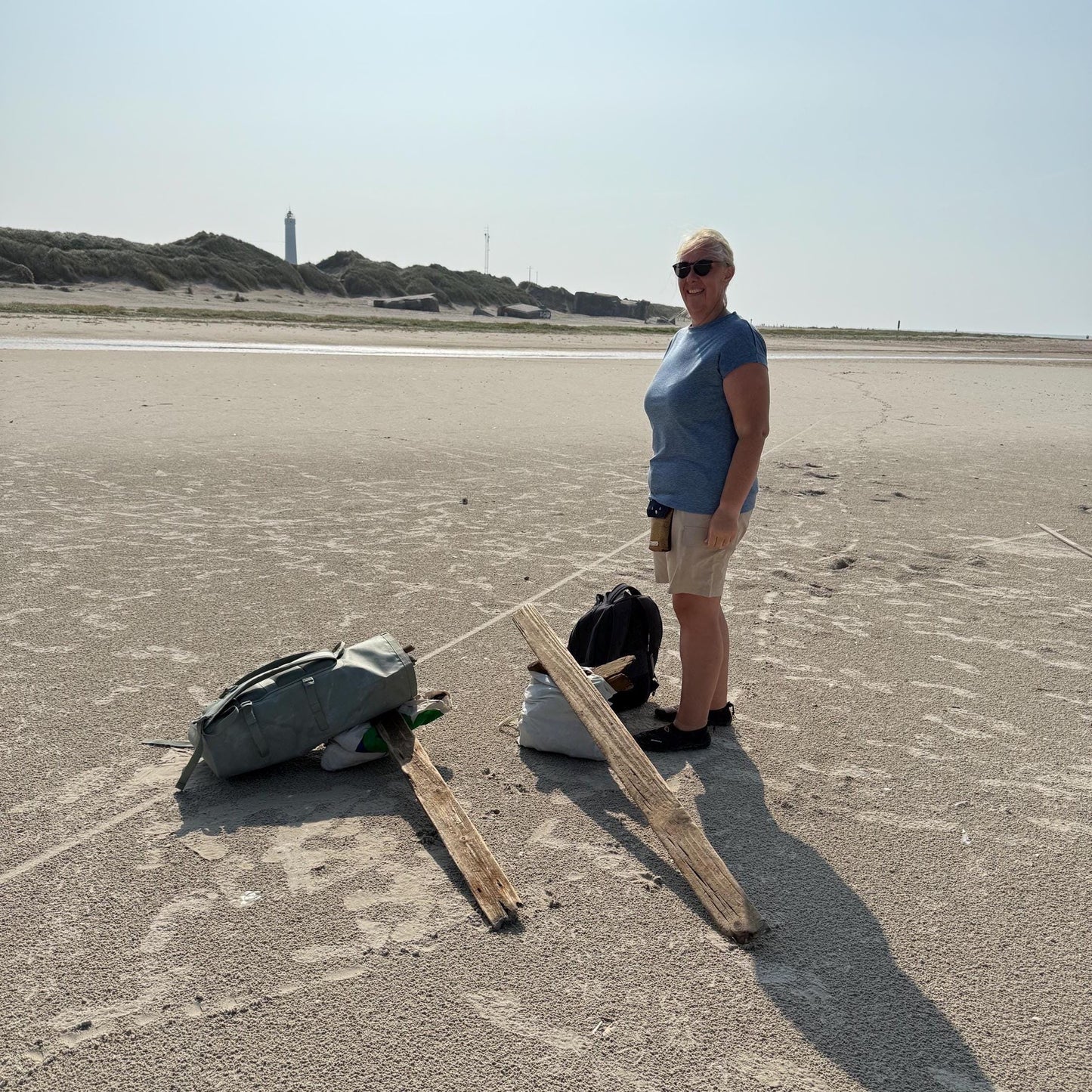Eine Person steht an einem Sandstrand und trägt ein blaues Oberteil, beige Shorts und eine Sonnenbrille.