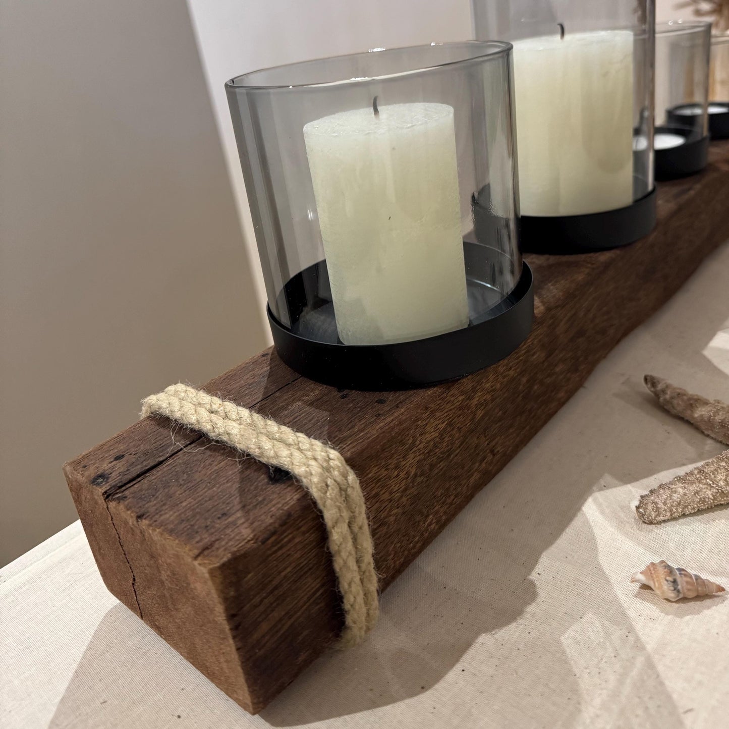 Ein rustikaler Kerzenhalter aus Holz mit vier weißen Säulenkerzen in Glas- und schwarzen Metallhaltern.