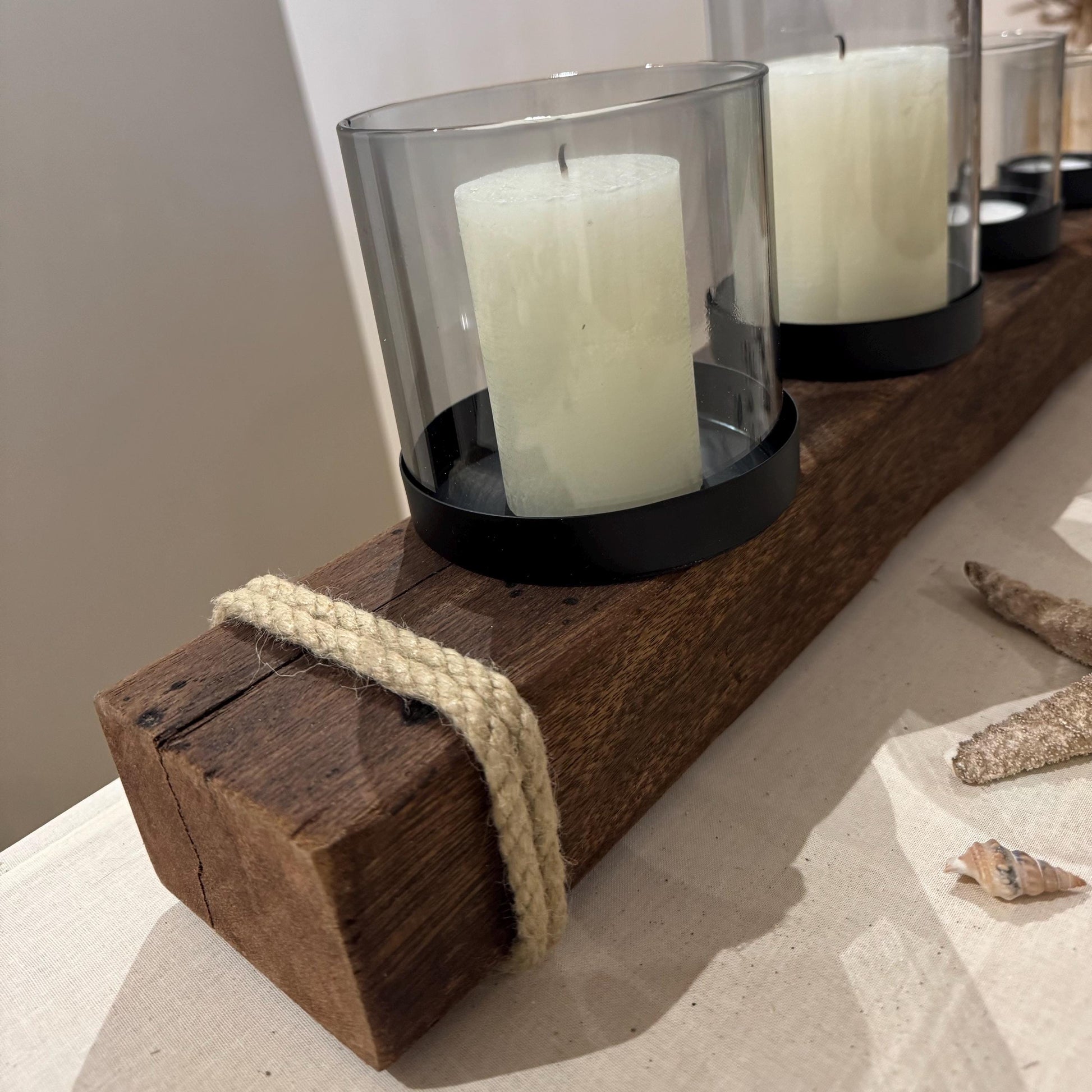 Ein rustikaler Kerzenhalter aus Holz mit vier weißen Säulenkerzen in Glas- und schwarzen Metallhaltern.
