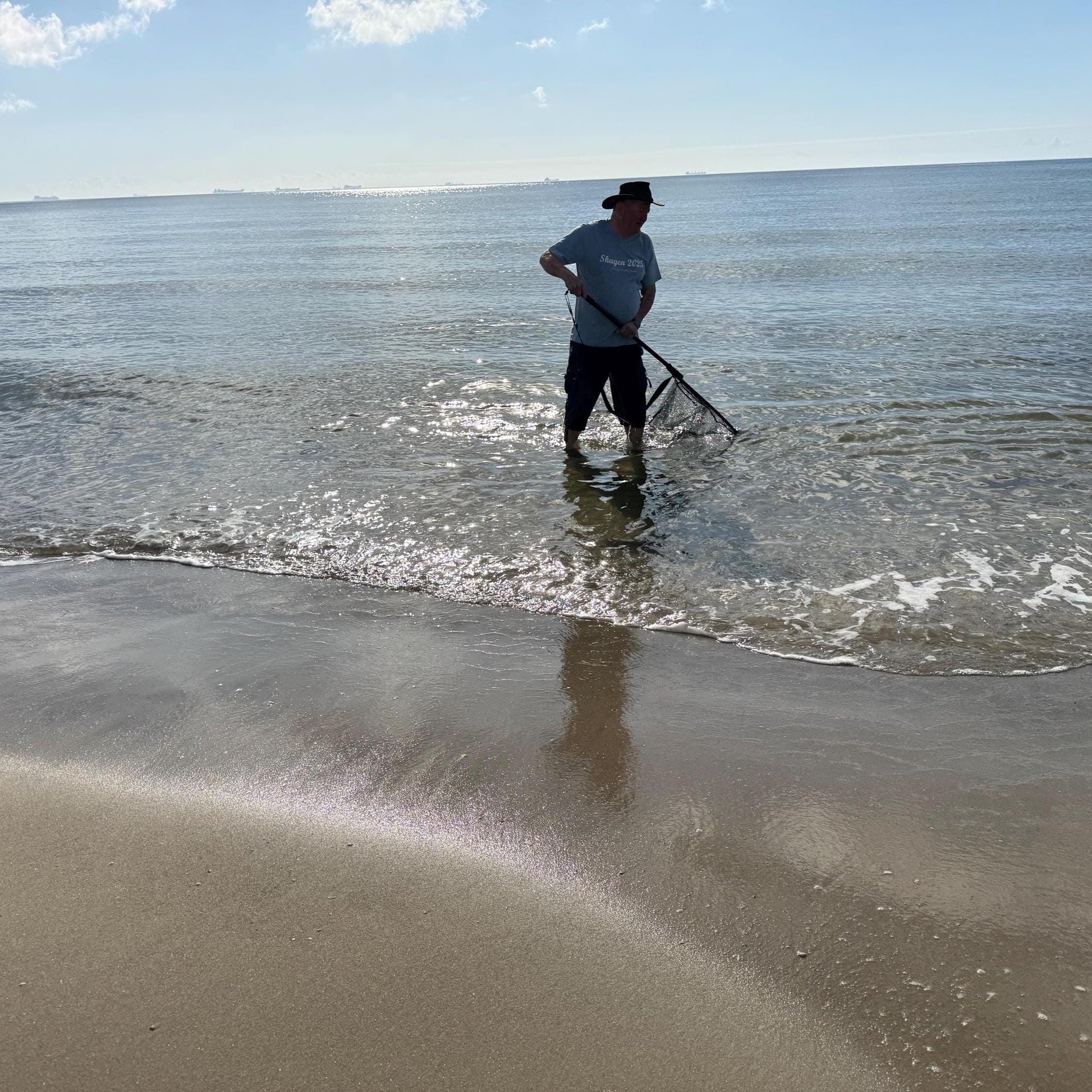 Eine Person steht im Meer mit einem Fischernetz, trägt einen Hut und ein hellblaues T-Shirt