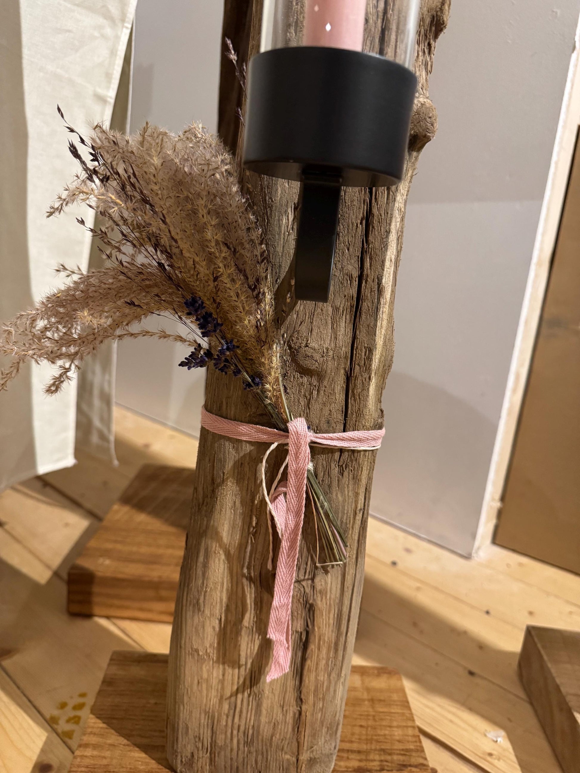 Hoher Treibholz-Kerzanständer mit Glaszylinder und brennender Stabkerze, dekoriert mit Trockenblumen und rosa Band