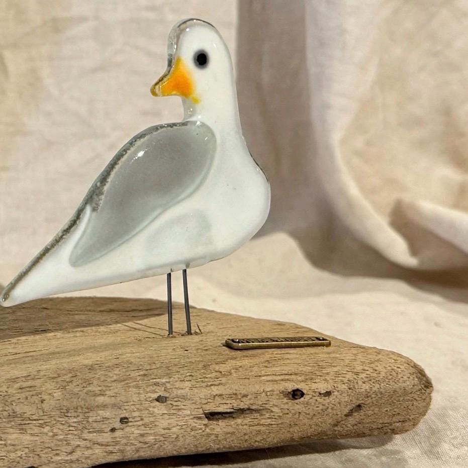 Detailansicht weiße Möwe aus Glas mit grauem Flügel und orangefarbenem Schnabel auf Treibholz stehend, maritime Glas Deko