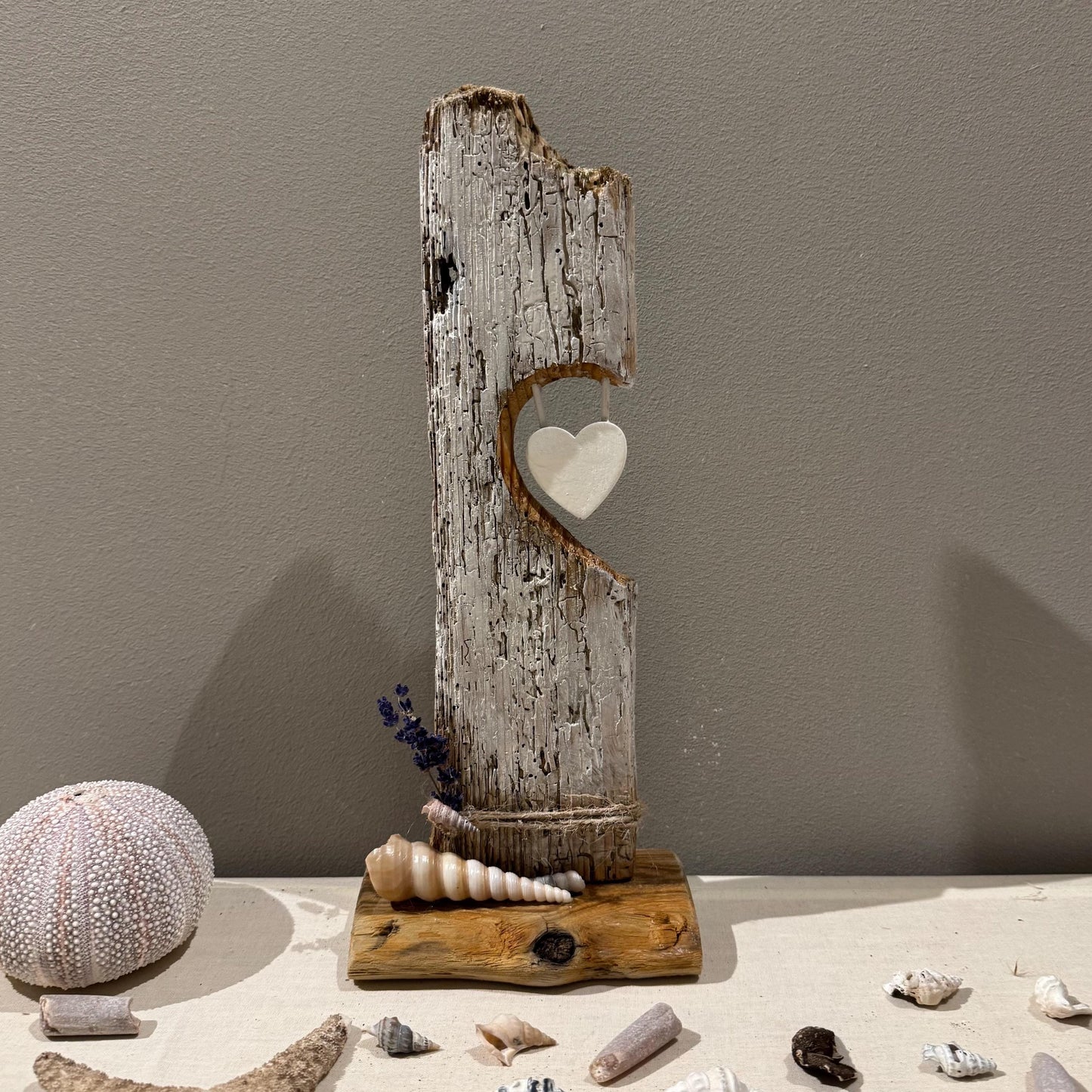 Eine verwitterte, grau gestrichene Holzskulptur mit herzförmigem Ausschnitt und einem hängenden weißen Herzen.